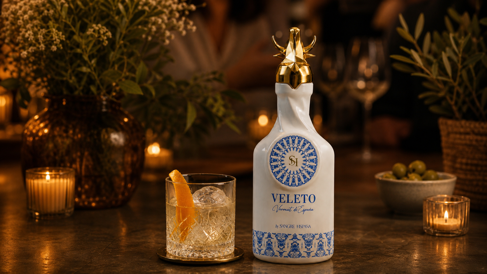Botella blanca del vermut Veleto de Sangre Hispana con motivo de azulejo azul y tapón dorado con cabeza de toro, junto a un cóctel